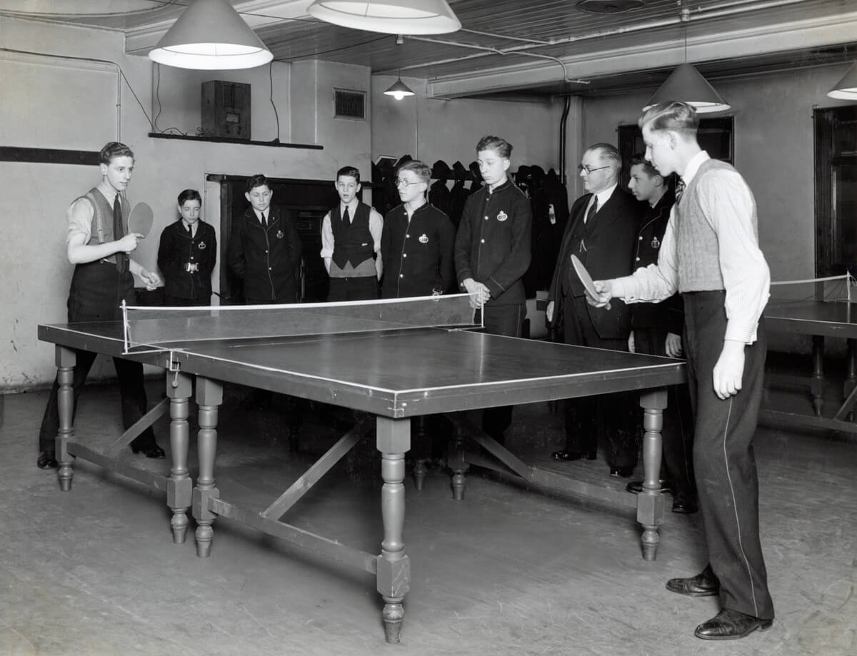 Two boys play table tennis while others watch. They are wearing their Telegram Messenger uniform.
