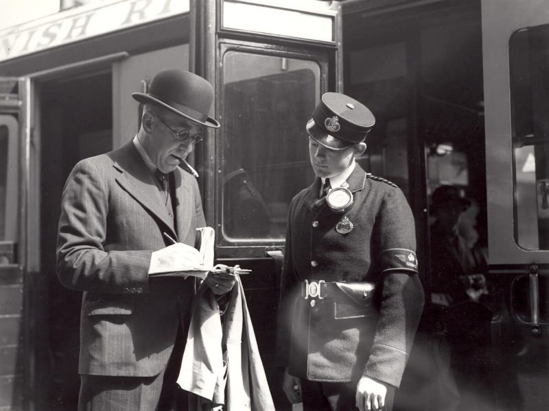 A young Telegram Messenger looks down at a telegram a man is writing on a paper form. The man is wearing a suit and hat and is smoking a cigar. Behind them is a train carriage with open doors.