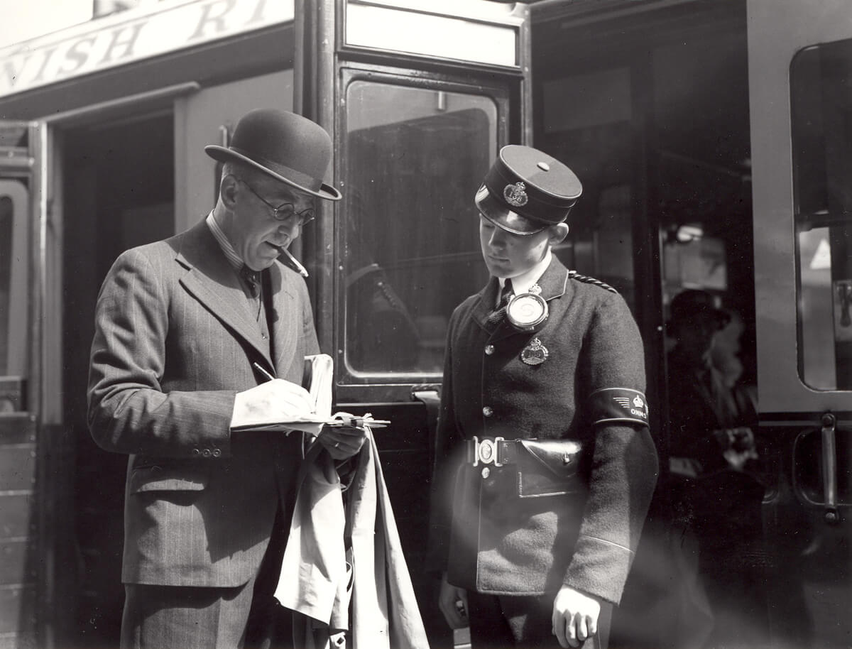 A young Telegram Messenger looks down at a telegram a man is writing on a paper form. The man is wearing a suit and hat and is smoking a cigar. Behind them is a train carriage with open doors.