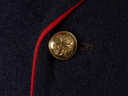 A brass button with a crown which top is similar to an upside down U. The crown is decorated with fleur de lys, a heraldic symbol of a lily flower with three petals, and a cross at the top and in the middle