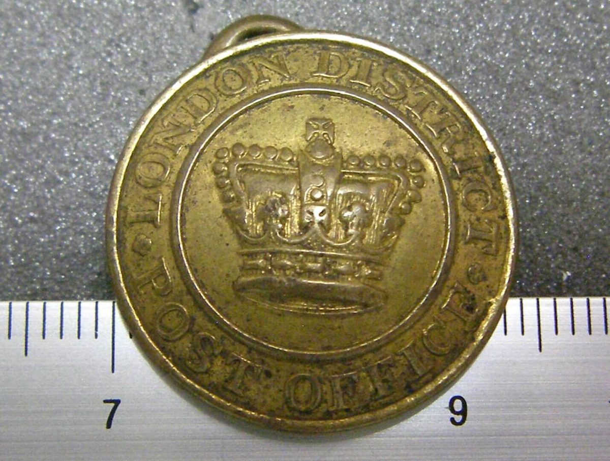 A brass bottom with a crown with flatten hearts-shaped arches, decorated with lily flowers and crosses at the top and middle areas. Around the crown, the text London District Post Office