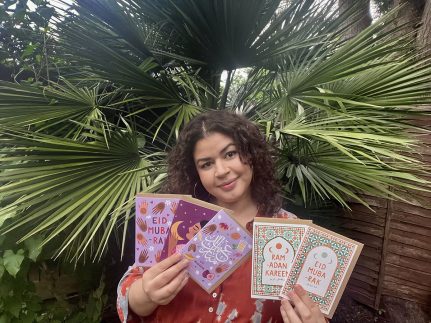 Sakina holds her cards and looks at the camera. She's outdoors and has a palm tree as background