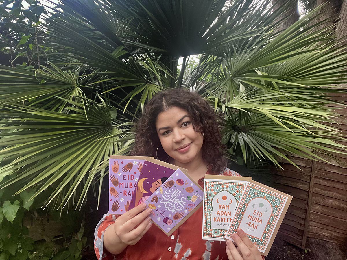 Sakina holds her cards and looks at the camera. She's outdoors and has a palm tree as background