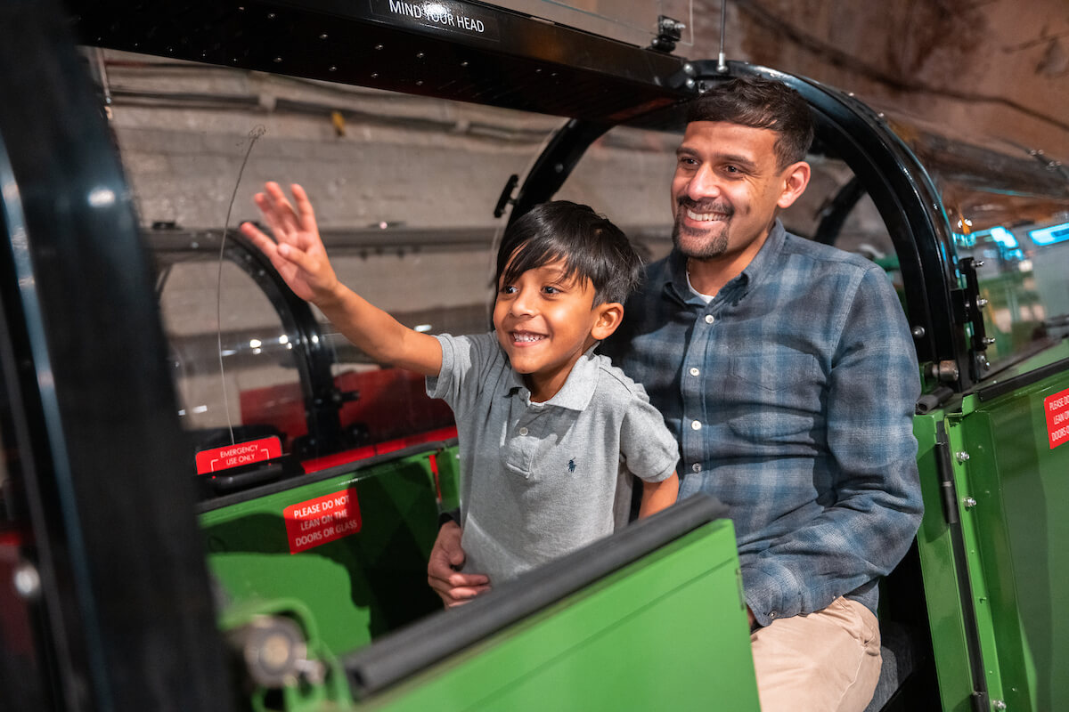 A parent and child riding a miniature green train.