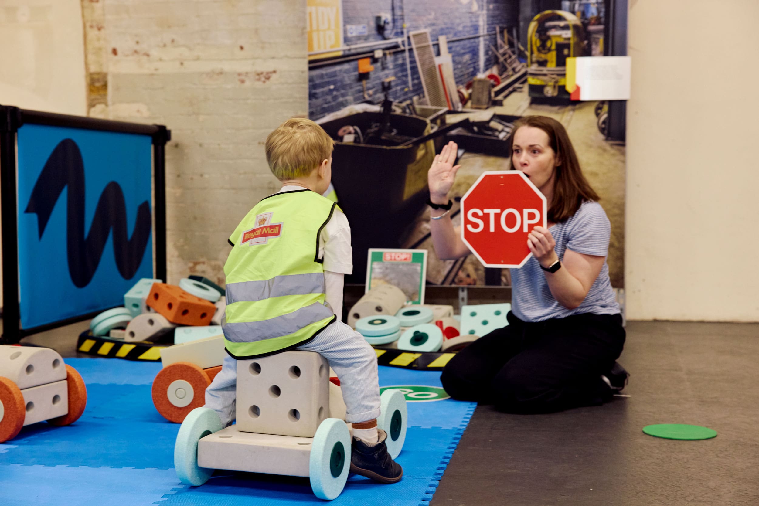 A little boy rides a car made of blocks. His mum sits by holding a stop sign.