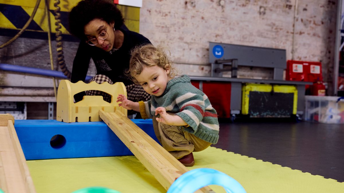 A little boy crouches by a ramp and watches his little toy car speed down it, through a colourful tunnel.