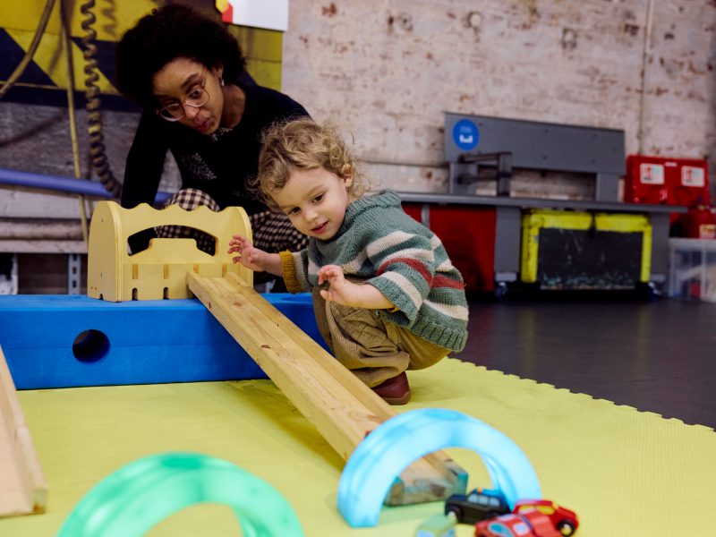 A little boy crouches by a ramp and watches his little toy car speed down it, through a colourful tunnel.