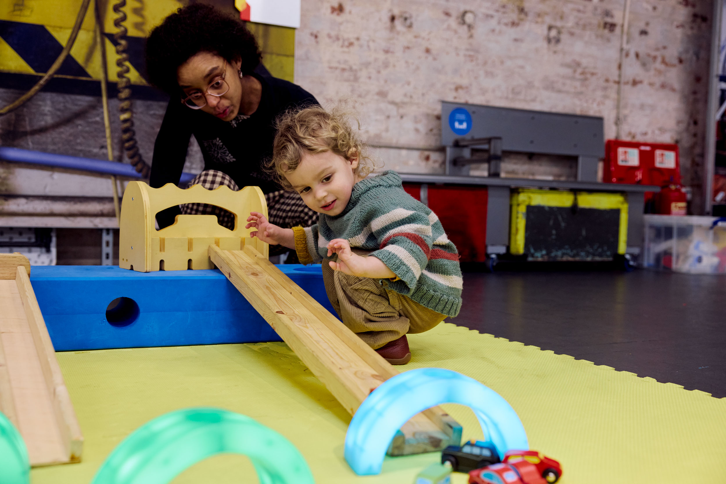 A little boy crouches by a ramp and watches his little toy car speed down it, through a colourful tunnel.