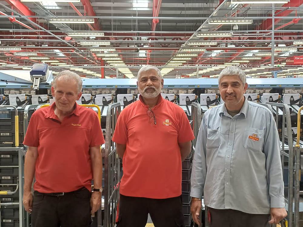 Three men in postal uniform, posing with mail centre equipment in the background.