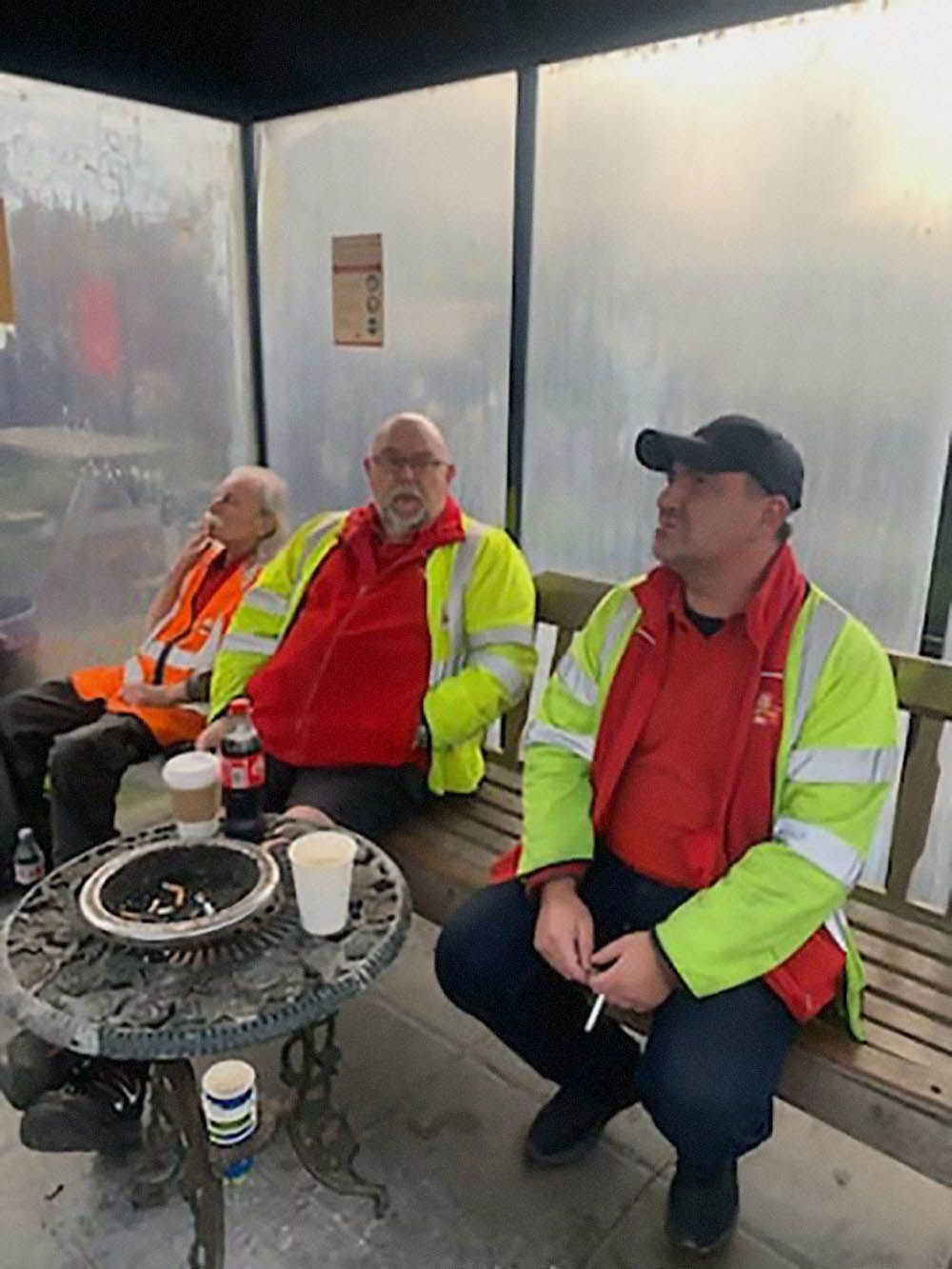Three men in postal uniform, sitting together at a bench with coffee cups.