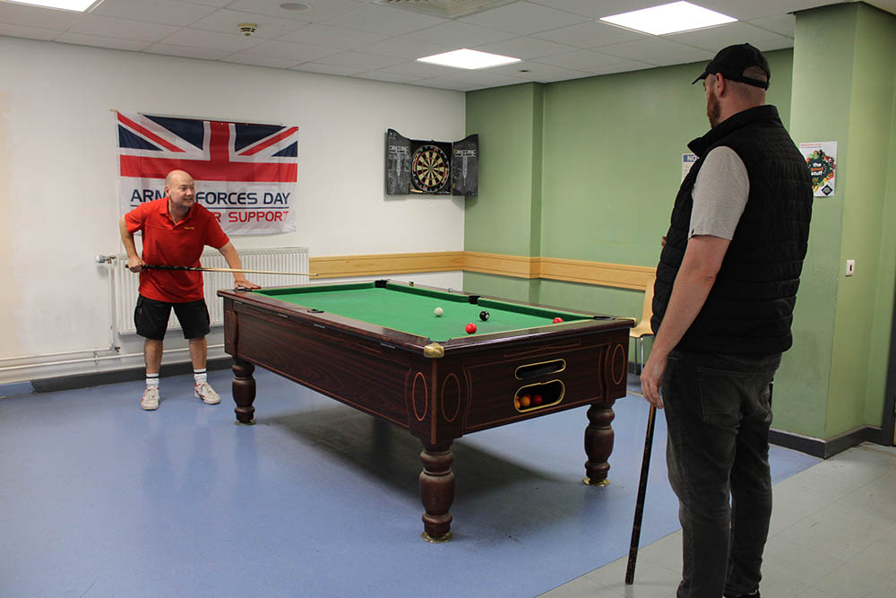 Two men playing snooker at the mail centre, with a banner commemorating Armed Forces Day in the background.