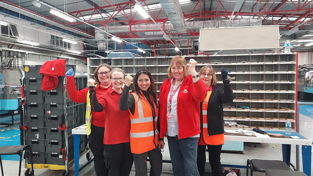 A group of postal workers in uniform at the mail centre, posing with sorting equipment in the background.