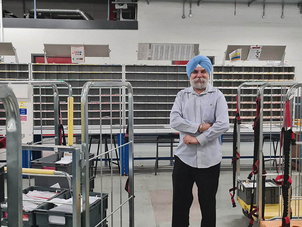 A man in postal uniform with trolleys and mail equipment in the background.