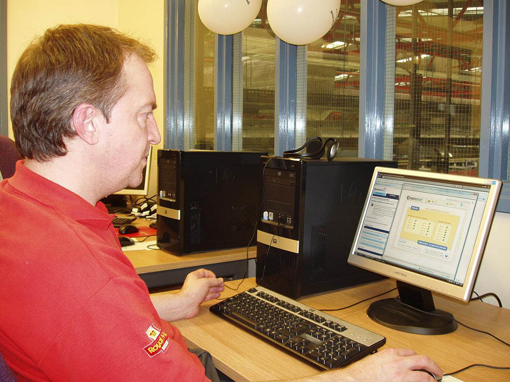 A man in red postal uniform working at a computer.