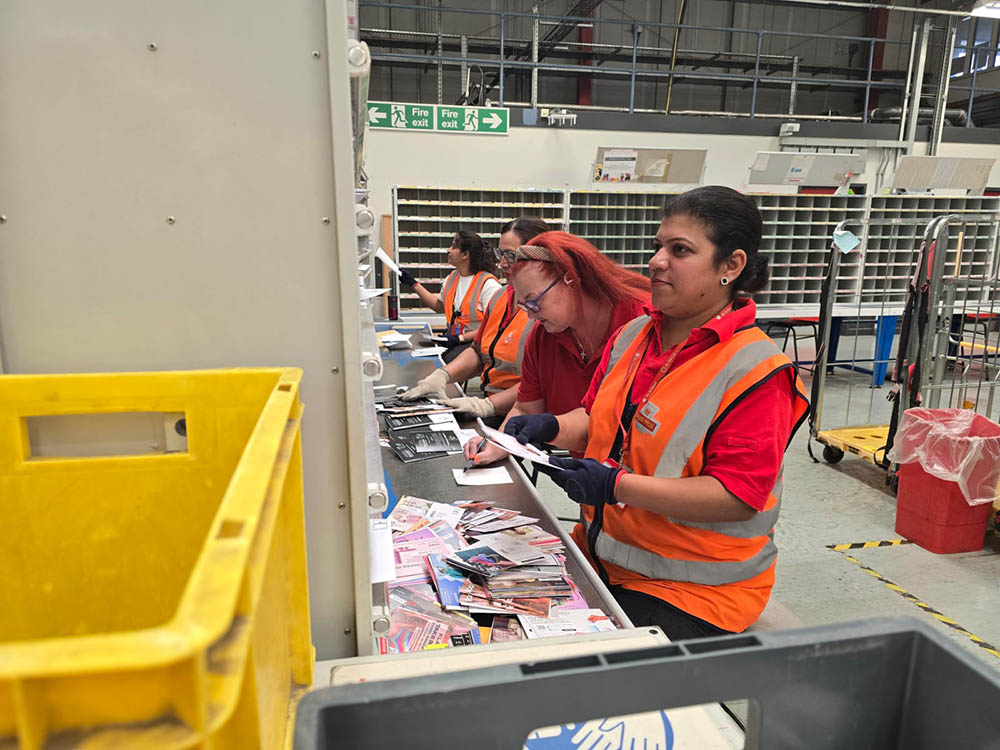 A row of postal workers in red postal uniforms, sorting letters.