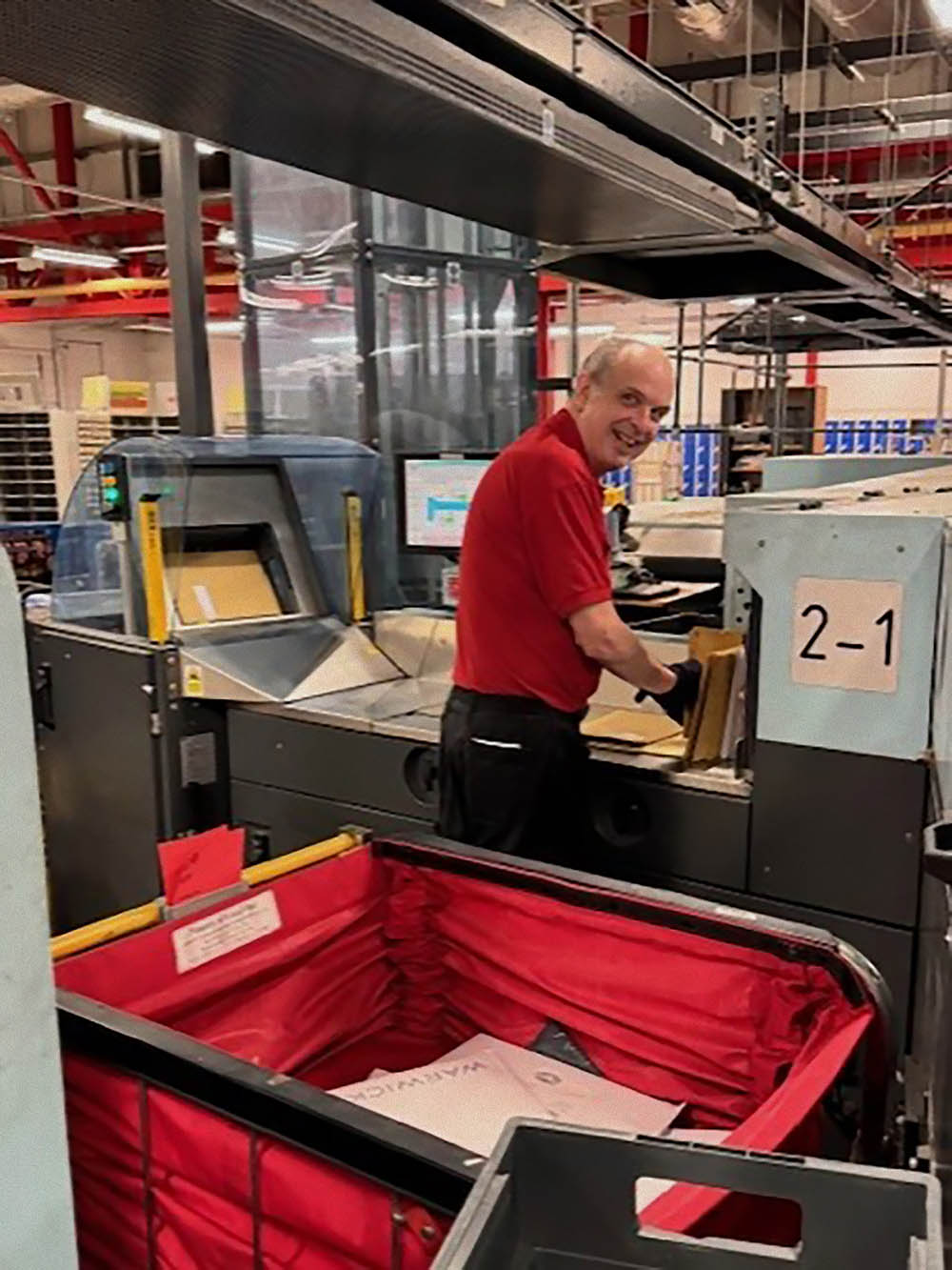 A man in a red postal uniform and trolley with sorting equipment.