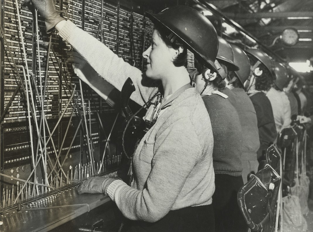Women telephonists operating a switchboard with steel helmets.