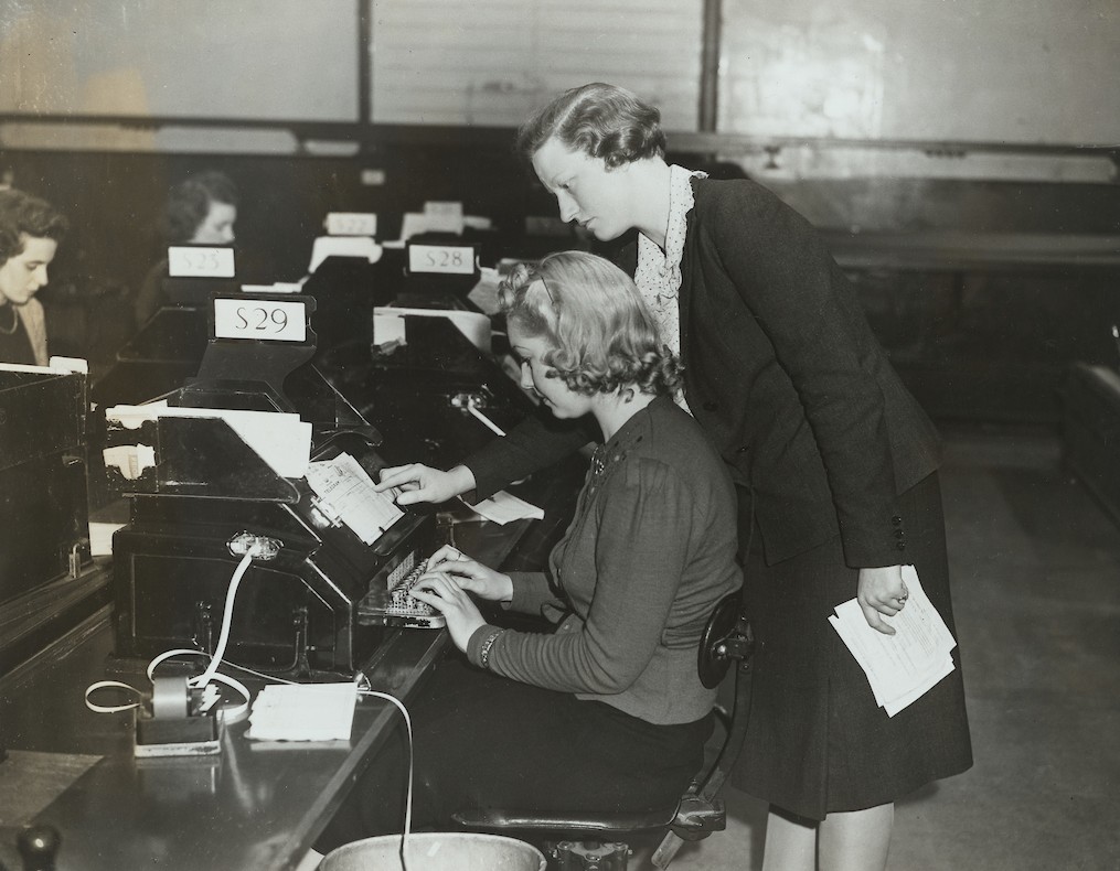 A woman telegraphist in training with a supervisor at a desk.